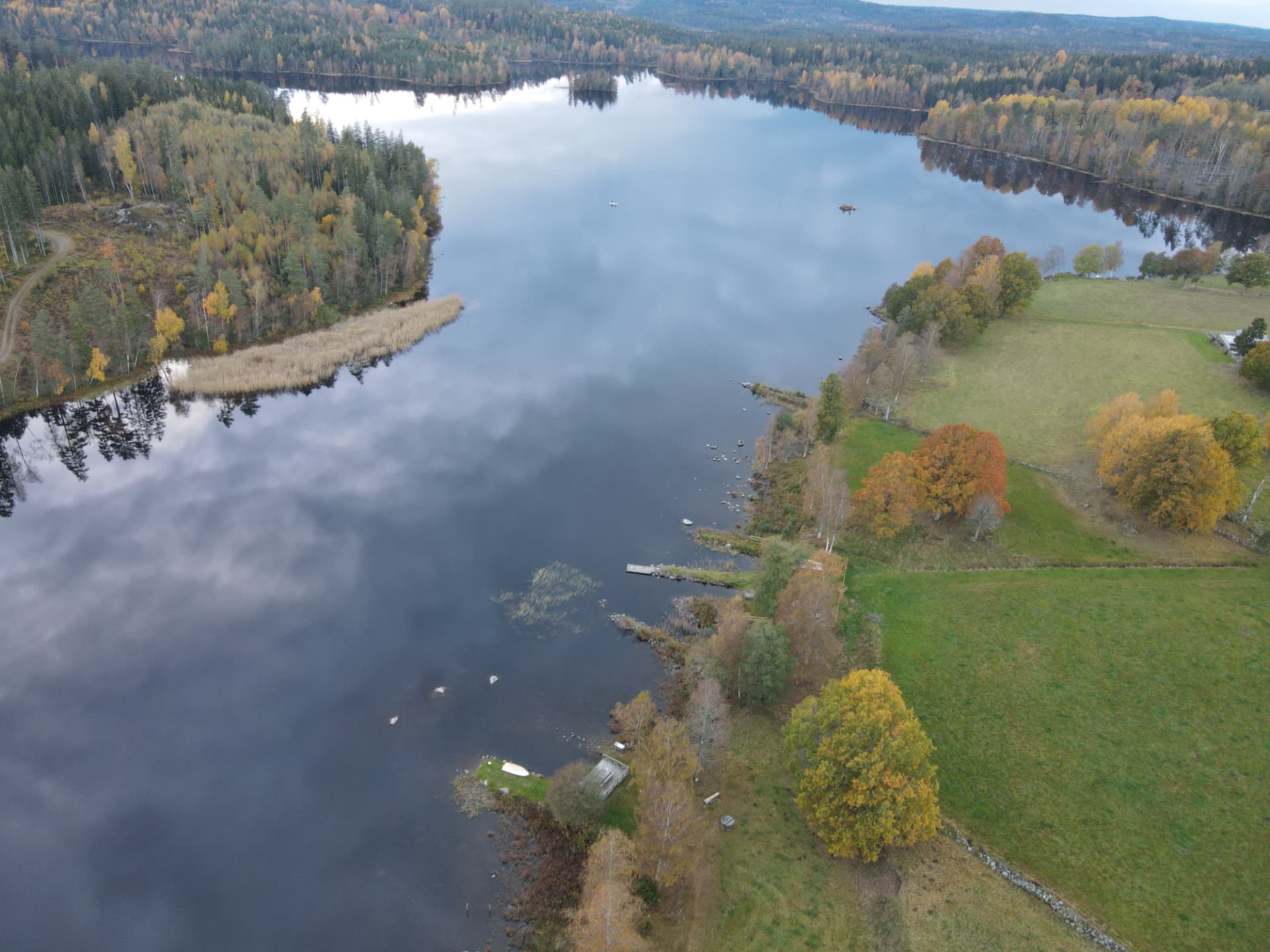 Luftfoto af en blank sø, der spejler skyerne, med enge og stengærder på bredden.
